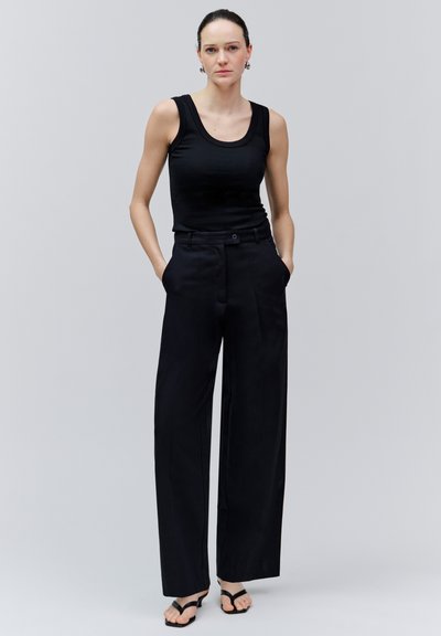 Femme aux cheveux foncés tirés en arrière porte un haut noir sans manches, un pantalon noir à jambes larges et des sandales noires, debout les mains dans les poches.