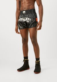 Schwarze Satin-Boxershorts mit elastischem Bund, ausgestattet mit weißem Branding und Seitentaschen; kombiniert mit schwarzen Sportschuhen.