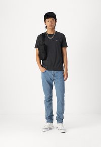 Grå kortermet t-skjorte med logo, blå denimjeans, hvite joggesko og en svart crossover-veske. Enkelt design og avslappet passform.