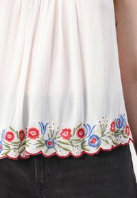 Blouse blanche texturée avec une bordure de broderie florale rouge, bleue et verte, portée sur un pantalon noir.