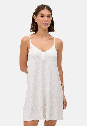 Mujer vistiendo un vestido tipo slip sin mangas, de color gris claro, hasta la rodilla, con detalles de encaje y finas tirantes, de pie frente a un fondo liso.