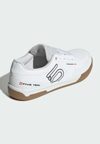 Scarpa sportiva bianca con suola in gomma marrone, superiore in materiale sintetico, dettagli perforati e accenti con logo. Presenta lacci tradizionali.