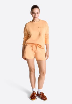 Tânără în picioare, purtând un top cu mânecă lungă de culoare piersică cu buzunar, pantaloni scurți asortați, șosete albe și adidași maro pe fundal alb.