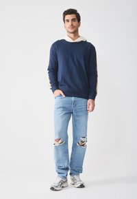 Felpa blu navy con cappuccio bianco, con strisce laterali beige. Jeans chiari in denim rovinato con ginocchia strappate. Sneakers grigie con accenti neri.