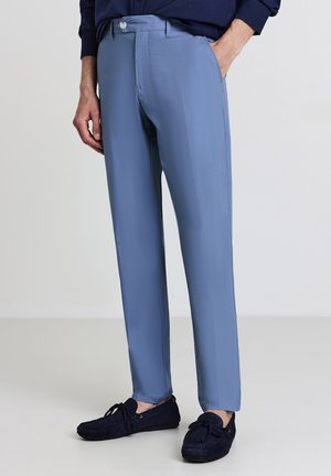Man wearing light blue slim-fit trousers with front pockets and dark blue suede loafers, standing against a plain light background.