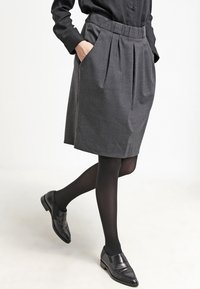 Jupe plissée grise avec une taille élastique, des poches latérales et une coupe au niveau des genoux, associée à des collants noirs et des chaussures.