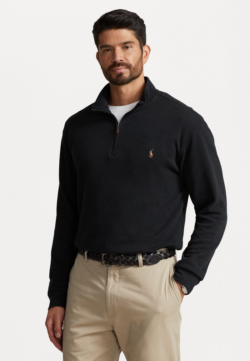 Black fleece quarter-zip pullover with ribbed cuffs, featuring a small embroidered logo on the chest. Styled with beige pants and a braided belt.