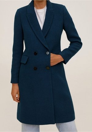 Osoba ubrana w ciemnoturkusowy dwurzędowy płaszcz z czarnymi guzikami, jedna ręka w kieszeni, w parze z jasnoniebieskimi dżinsami i białą koszulą.