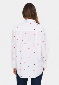 White long-sleeved shirt with red heart patterns, featuring a button-down collar and a curved hem. Soft fabric with a relaxed fit.