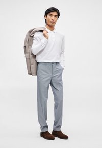 Λευκό πουκάμισο με μακριά μανίκια, ανοιχτό γκρι σακάκι περασμένο στον ώμο, μπλε καρό παντελόνι και καφέ σουέτ μπότες. Μινιμαλιστικό φόντο.