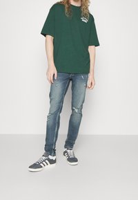 Camiseta de algodón verde con logo, combinada con unos vaqueros claros desgastados. El calzado presenta zapatillas grises y blancas con acentos a rayas.