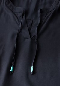 Zwarte tuniek met een V-hals en striksluiting. Kenmerkt zich door tealkleurige accenten op de uiteinden van de strik. Glad, lichtgewicht materiaal met een subtiele glans.