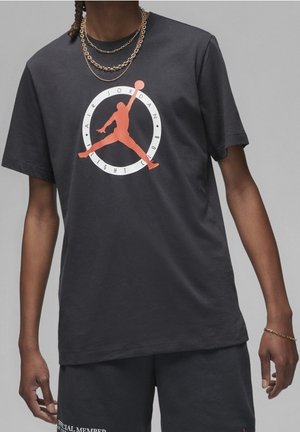 Osoba nosząca ciemnoszarą koszulkę Air Jordan z czerwonym logo i pasujące spodenki, uzupełnione złotymi łańcuszkami i bransoletką.