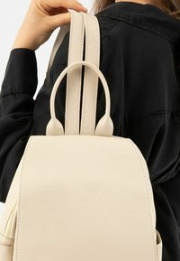 Beżowy skórzany plecak z dwoma paskami na ramiona i górnym uchwytem. Charakteryzuje się gładką fakturą oraz kieszenią na suwak z boku.