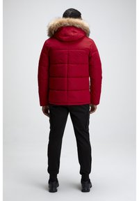 Veste rouge matelassée avec capuche doublée de fourrure, présentant des coutures horizontales. Portée avec un pantalon cargo noir et des baskets noires.