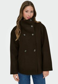 Abrigo marrón de doble botonadura con un alto cuello, grandes botones y bolsillos laterales. La tela parece suave con una textura cálida.