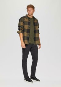 Flanellhemd im Schwarz-Olivgrün-Karomuster, kombiniert mit einem olivgrünen T-Shirt, schwarzen Jeans und schwarzen Sneakern.