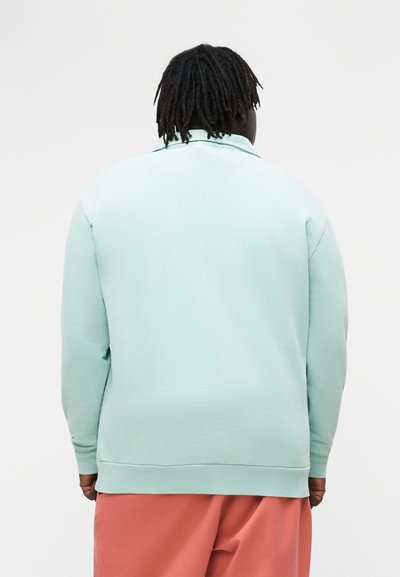 Oseba s temnimi dredloki nosi svetlo mintno zelen pulover in koralno rožnate hlače, obračena stran proti preprosti podlagi.