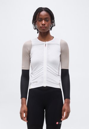 Naine, kes kannab valget lühikeste varrukatega lukuga rattasärki, millel on beežid ja mustad pikad varrukad ning mustad rattapüksid, seisab valge tausta ees.