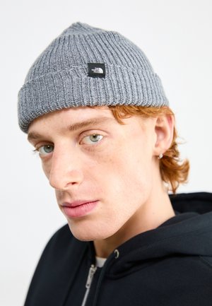 Graue Strickmütze mit strukturiertem Muster und einem kleinen schwarzen Logopatch. Die Mütze sitzt eng und verjüngt sich leicht nach oben.