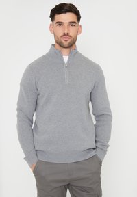 Grijze pullover trui met een halve rits kraag, geribde manchetten en zoom. Zachte stoftextuur, aangesloten ontwerp. Draagt over een wit shirt.