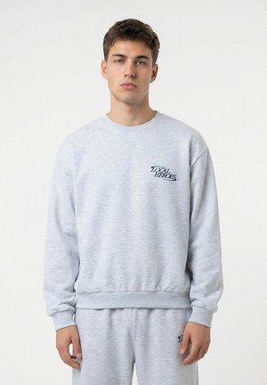 Młody mężczyzna w jasnoszarej bluzie i pasujących spodniach dresowych z logo "Local Heroes" na piersi i udzie, stojący na białym tle.