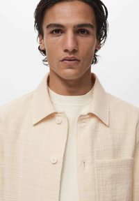Light beige shirt with a textured fabric, open collar, and buttons. Worn over a cream sweater, highlighting a layered style.