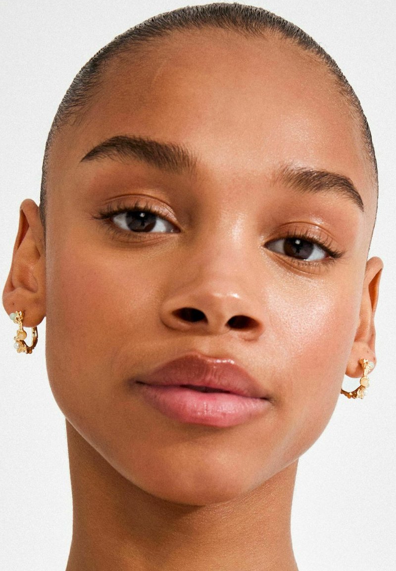 Gold hoop earrings with textured accents, featuring small decorative elements and curvy shape, set against smooth skin.