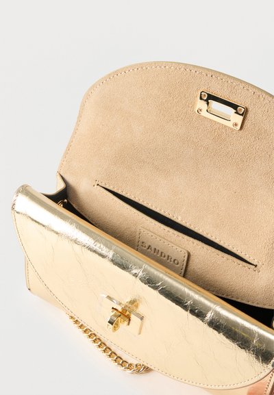 Μικρή μεταλλική χρυσή δερμάτινη τσάντα με σουέντ εσωτερικό, χρυσό κούμπωμα και λουρί αλυσίδα, ανοιχτή για να δείξει εσωτερική τσέπη και την ετικέτα της μάρκας Sandro.