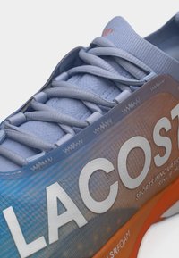 Gros plan d'une chaussure de sport bleue et orange avec des lacets épais et un grand texte blanc "LACOSTE" sur le côté.