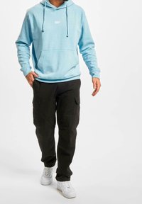 Felpa leggera blu con cappuccio e cordini, abbinata a pantaloni cargo neri e sneaker bianche. Design semplice, vestibilità casual.