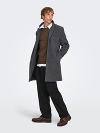 Manteau en laine gris avec un col cranté, poche avant, porté par-dessus un pull marron et une chemise blanche, associé à un pantalon noir et des chaussures marron.