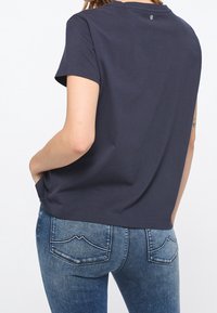 Kvinna som bär en marinblå kortärmad skjorta och blå denimjeans, stående med händerna i fickorna, vänd bort från kameran.