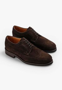 Zapatos de vestir de ante marrón con perforaciones decorativas, cierre de cordones y suela de goma plana. Punta ligeramente redondeada con forro interior suave.