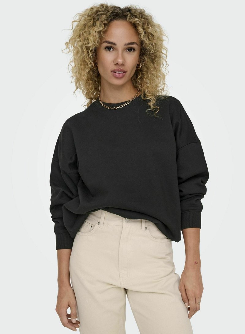 Svart oversized sweatshirt med ribbade ärmslut, rundad halsringning och mjukt material, ihop med ljusbeige hög midja byxor. Enkel guldkedja som accent.