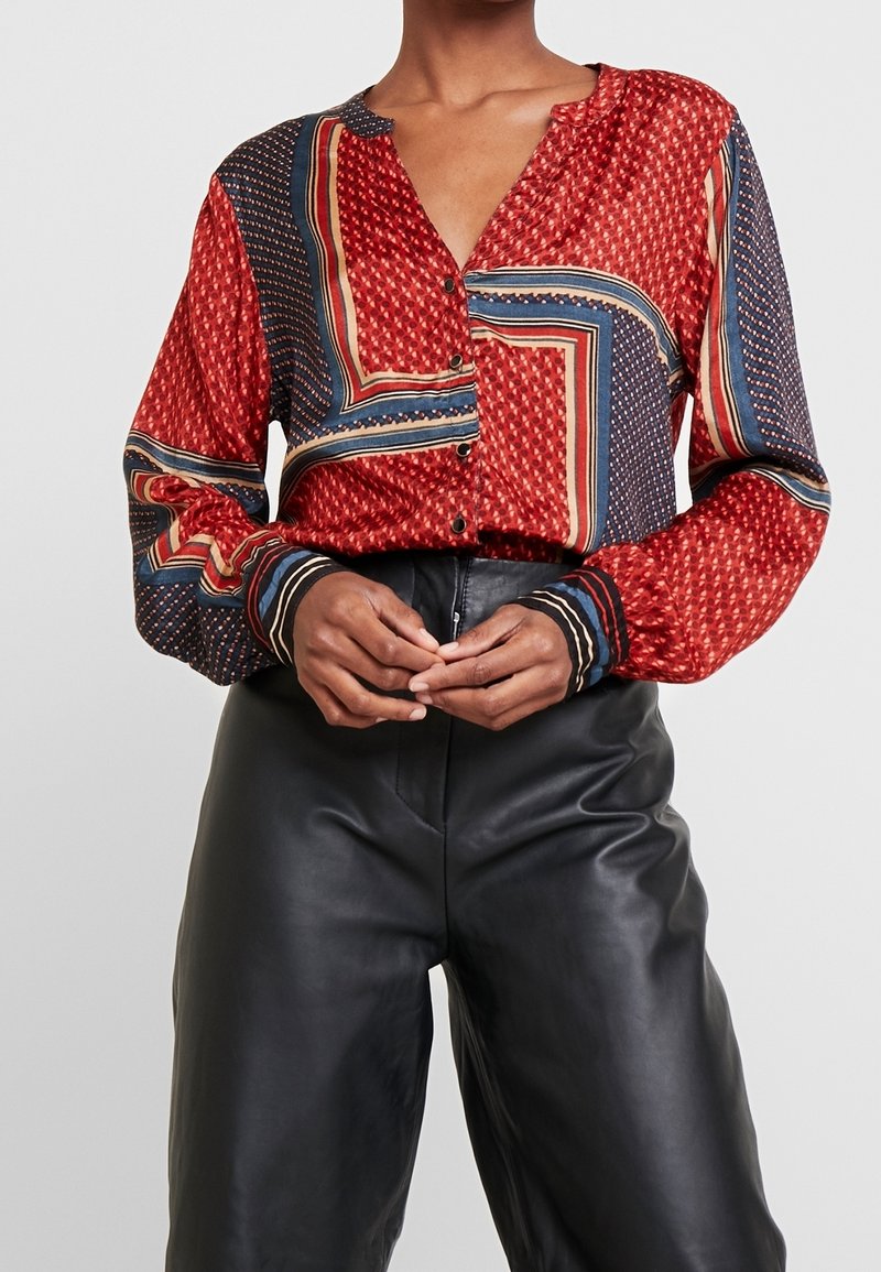 Femme portant une chemise rouge à motifs à manches longues boutonnée rentrée dans un pantalon en cuir noir taille haute, debout, les mains jointes.