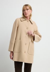 Beige jas met een grote kraag, vijf knopen en twee voorkruiszakken. Heeft gestructureerde mouwen. Eenvoudig, gestructureerd ontwerp.