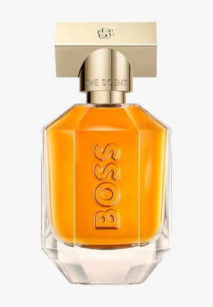 Przezroczysta, wielościenna butelka perfum ze szkła z bursztynowym płynem, złotą nakrętką z wygrawerowanym napisem "THE SCENT" oraz wytłoczonym napisem "BOSS" z przodu.