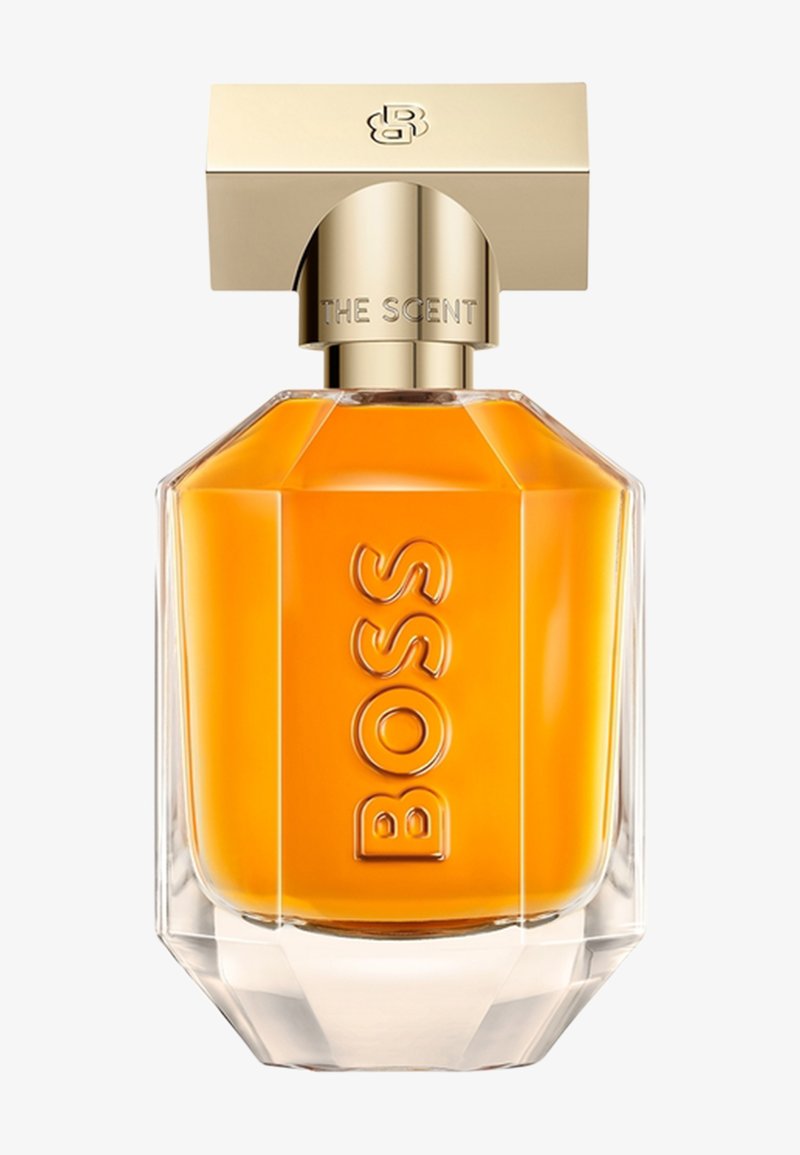 Flacon de parfum en verre clair facetté avec un liquide ambré, bouchon doré gravé "THE SCENT" et "BOSS" en relief sur le devant.