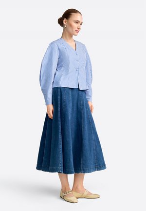 Vrouw met een blauw-wit gestreepte blouse met lange mouwen, een donkerblauwe A-lijn midi spijkerrok, beige ballerina's en zilveren oorbellen, staand met de handen langs het lichaam.