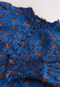 Blouse bleue à motifs avec patte de boutonnage, bordure en dentelle, col froufrou et poignets smockés, posée à plat sur une surface claire.