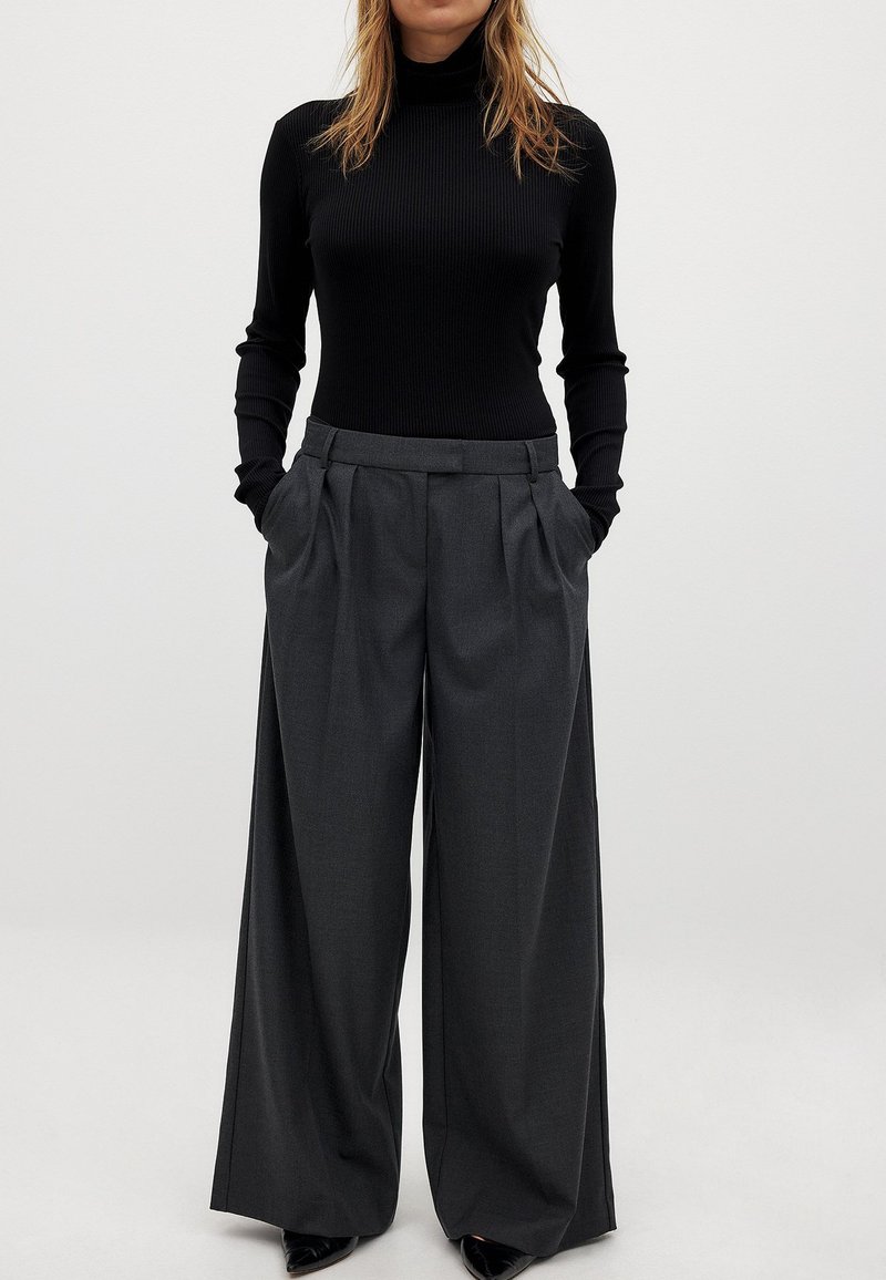 Zwarte geribbelde coltrui en grijze wijde pantalon met plooien, met een gladde textuur en zijzakken, gecombineerd met zwarte hakken.