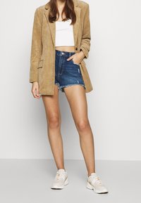 Mujer vistiendo un blazer de pana marrón claro, una camiseta corta blanca, pantalones cortos de mezclilla desgastados y zapatillas blancas, de pie contra un fondo liso.