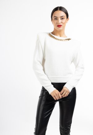 Vrouw draagt een witte trui met gouden kettingdetail en zwarte leren broek, staande met gevouwen handen voor zich tegen een witte achtergrond.