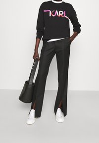 Sudadera negra con el logo rosa "KARL", combinado con pantalones anchos negros con aberturas laterales y zapatillas blancas. Accesorio: bolso tipo bucket negro.