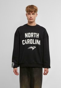 Svart hettegenser med "NORTH CAROLINA" i fet hvit tekst og et logo. Har droppe skuldre og en ribbestrikket hem. Ledig passform.