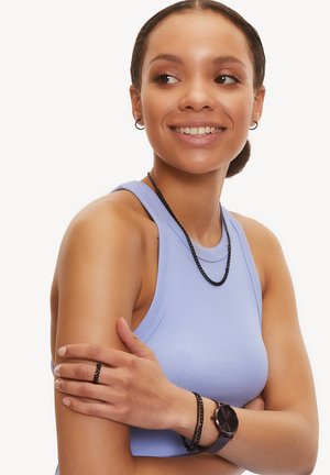 Les bijoux noirs incluent un collier superposé, un bracelet et une bague. La personne porte un débardeur bleu clair et une montre noire à son poignet.