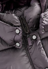 Geacă puffer de culoare mov închis cu o textură netedă și strălucitoare. Are nasturi metalici argintii și un fermoar central cu un tab său negru.