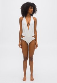 Kvinde med krøllet hår iført en hvid badedragt med dyb V-udskæring og halterneck, stående barfodet mod en ensfarvet hvid baggrund.