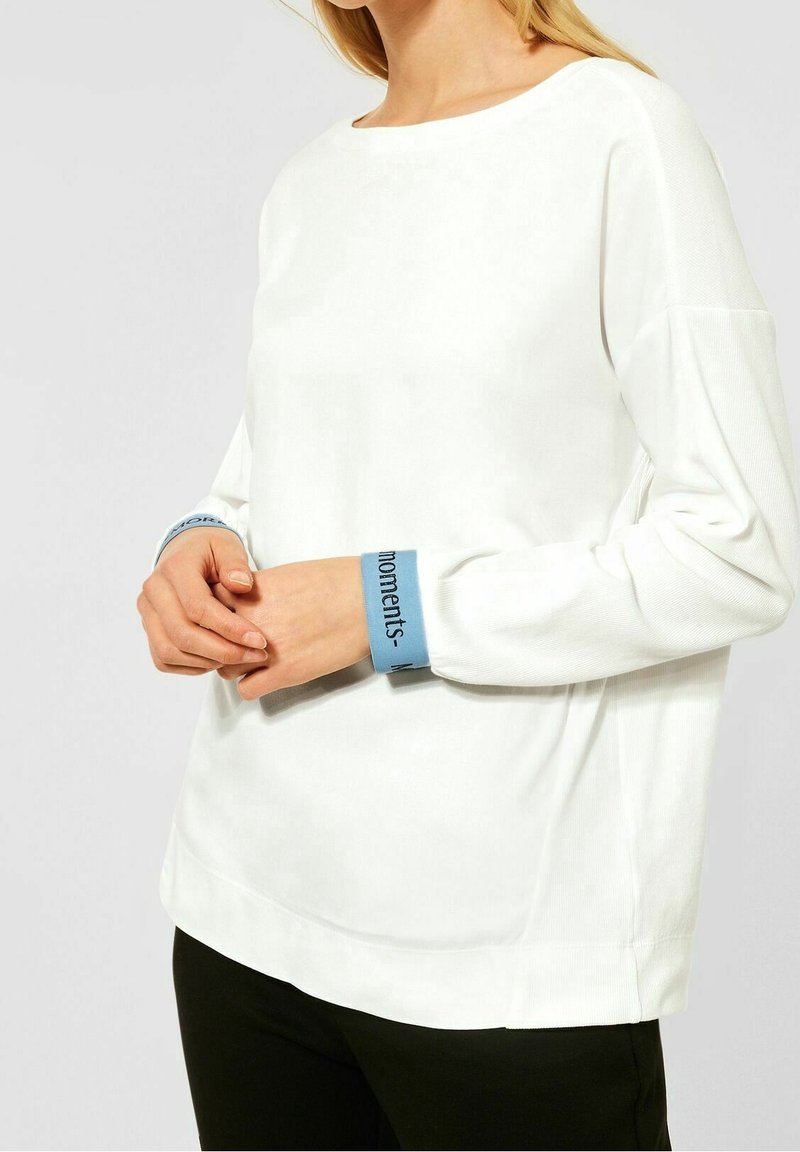 Camiseta blanca de manga larga con cuello redondo y puños azules con texto. Confeccionada en material suave, tiene un corte holgado y una textura suave.